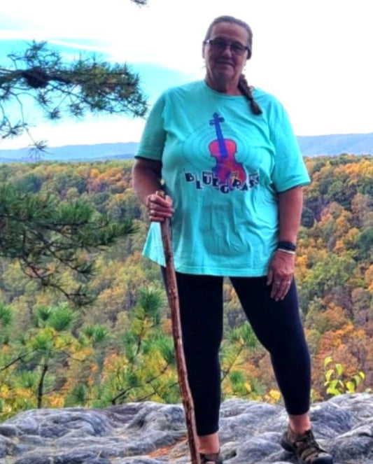 Person standing on a rocky outcrop wearing a fiddle t-shirt with the words bluegrass with a scenic view of trees in the background