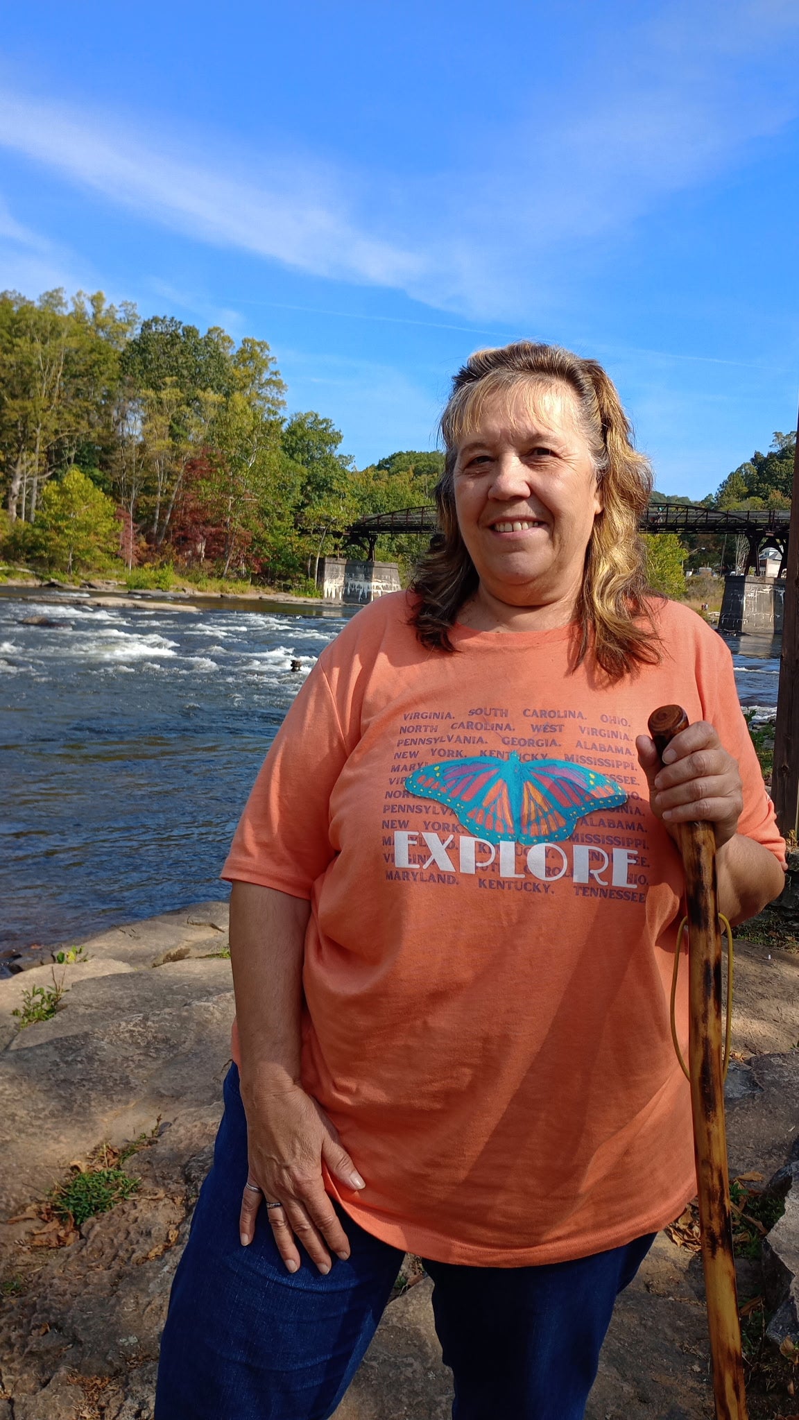 Woman standing by a river with trees and a bridge in the background, wearing an orange 'EXPLORE' t-shirt.
