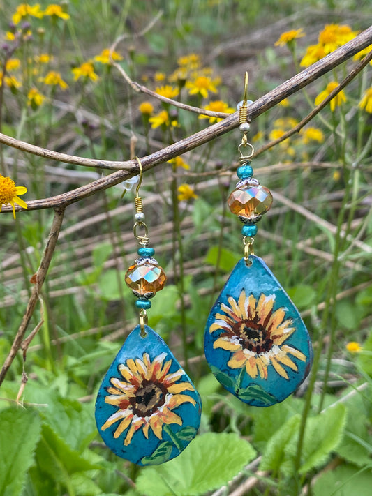 Teardrop-shaped earrings with sunflower design hanging on a branch with green leaves and yellow flowers in the background.