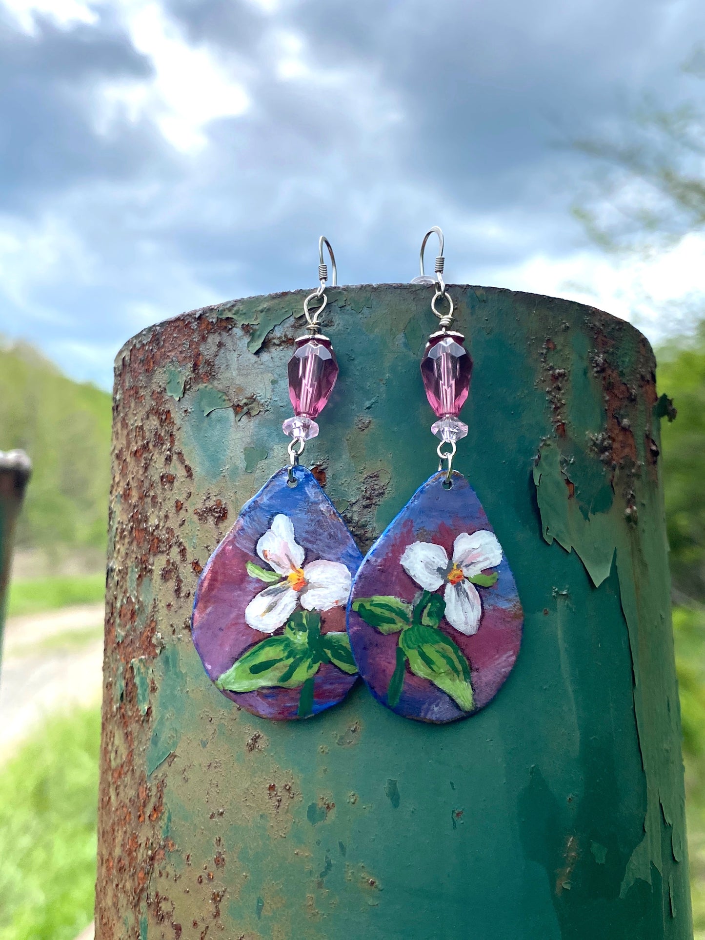 Teardrop-shaped earrings with floral design hanging on a rusty metal surface outdoors.