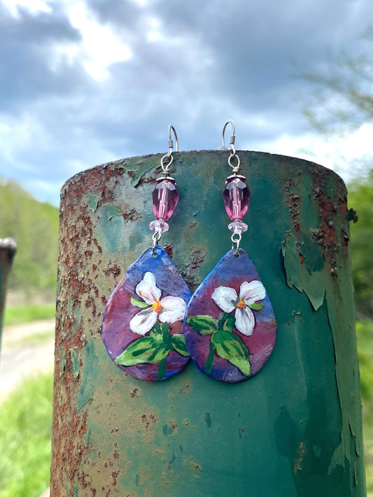 Teardrop-shaped earrings with floral design hanging on a rusty metal surface outdoors.
