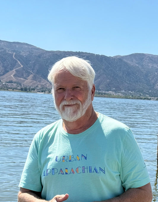 Man with white hair and beard wearing a light blue 'Urban Appalachian' shirt standing by a lake with mountains in the background.