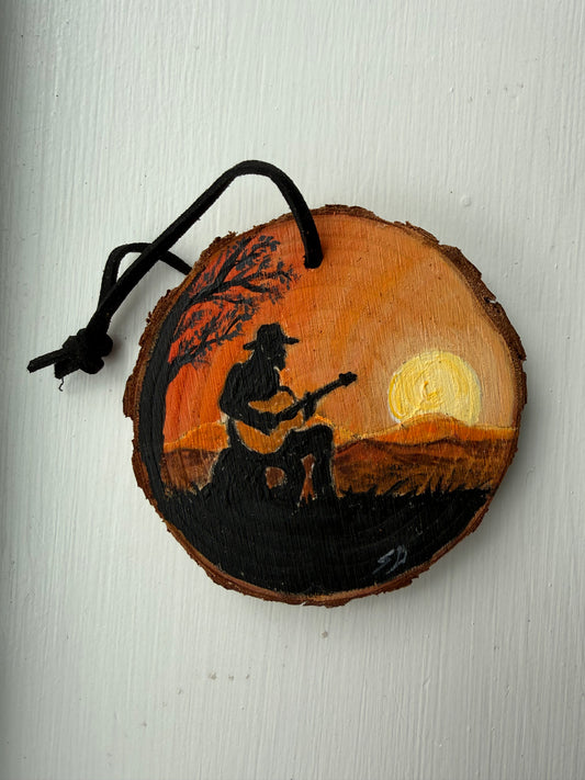 Wooden ornament with a painted scene of a person playing guitar under a moonlit sky