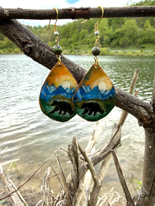 Pair of earrings with bear design hanging on a branch by a lake