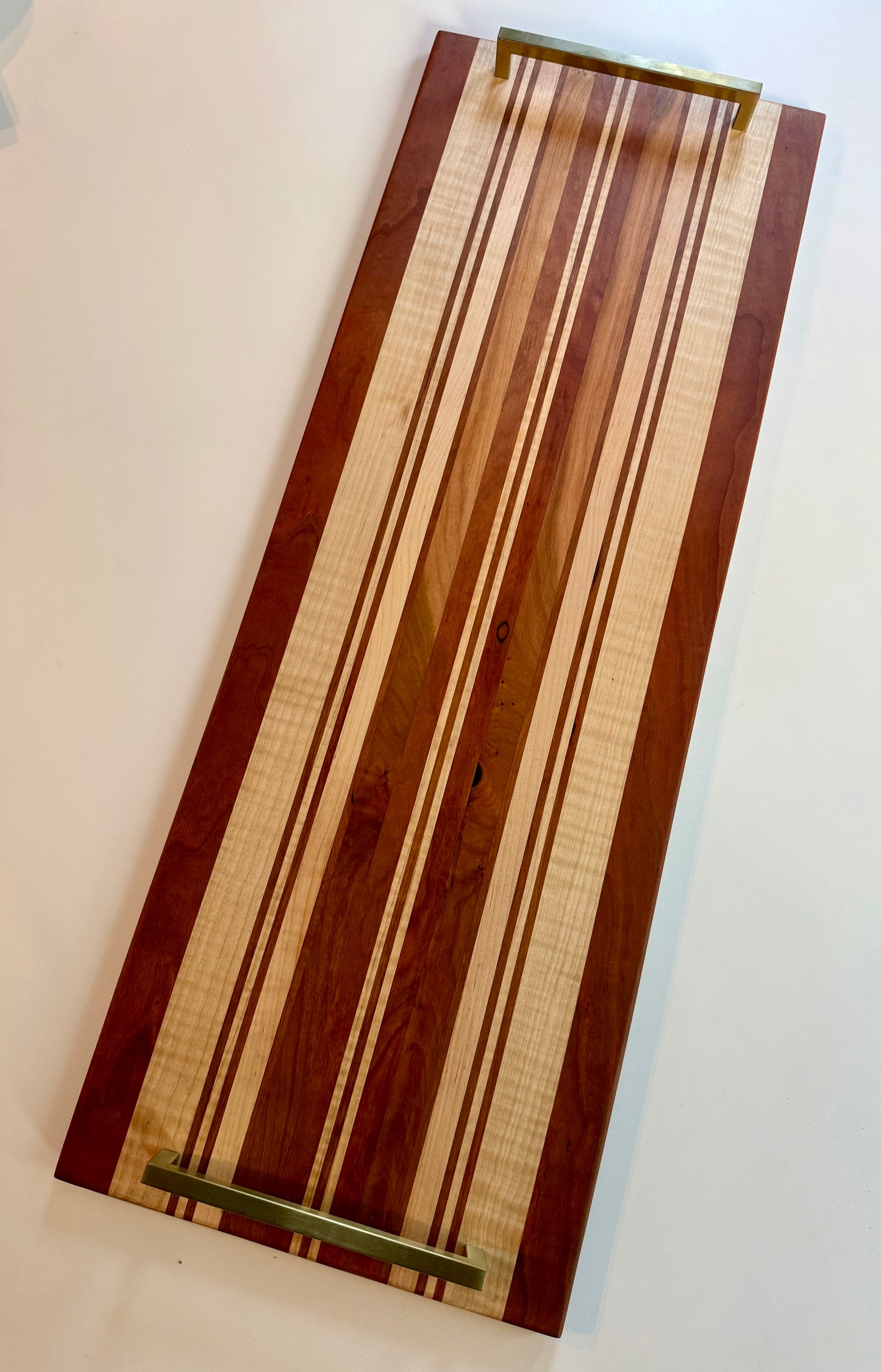 A rectangular wooden serving board with a striped pattern of light and dark wood tones, featuring brass colored handles