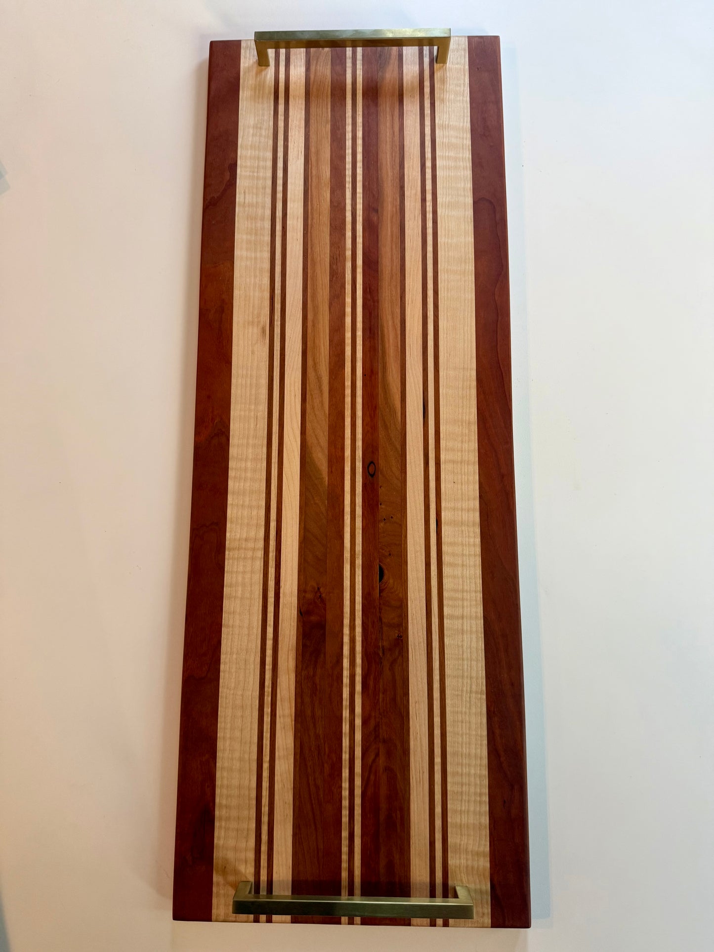 A rectangular wooden serving board with a striped pattern of light and dark wood tones, featuring brass colored handles