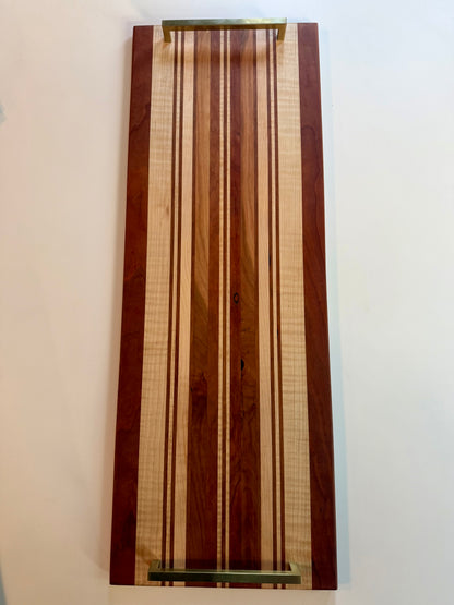 A rectangular wooden serving board with a striped pattern of light and dark wood tones, featuring brass colored handles