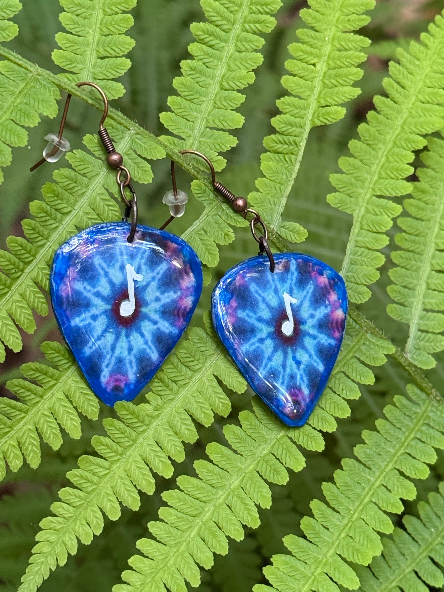 Blue tie dye teardrop-shaped earrings with a music note design on on green fern leaves