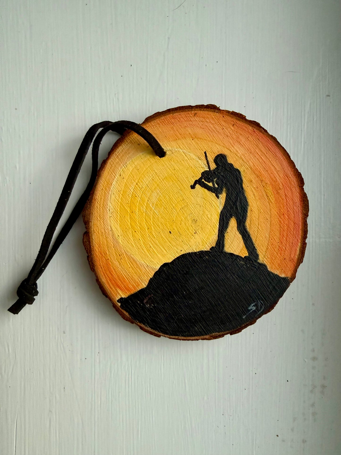 Wooden ornament with a silhouette of a person playing a fiddle on a rock against a sunset sky