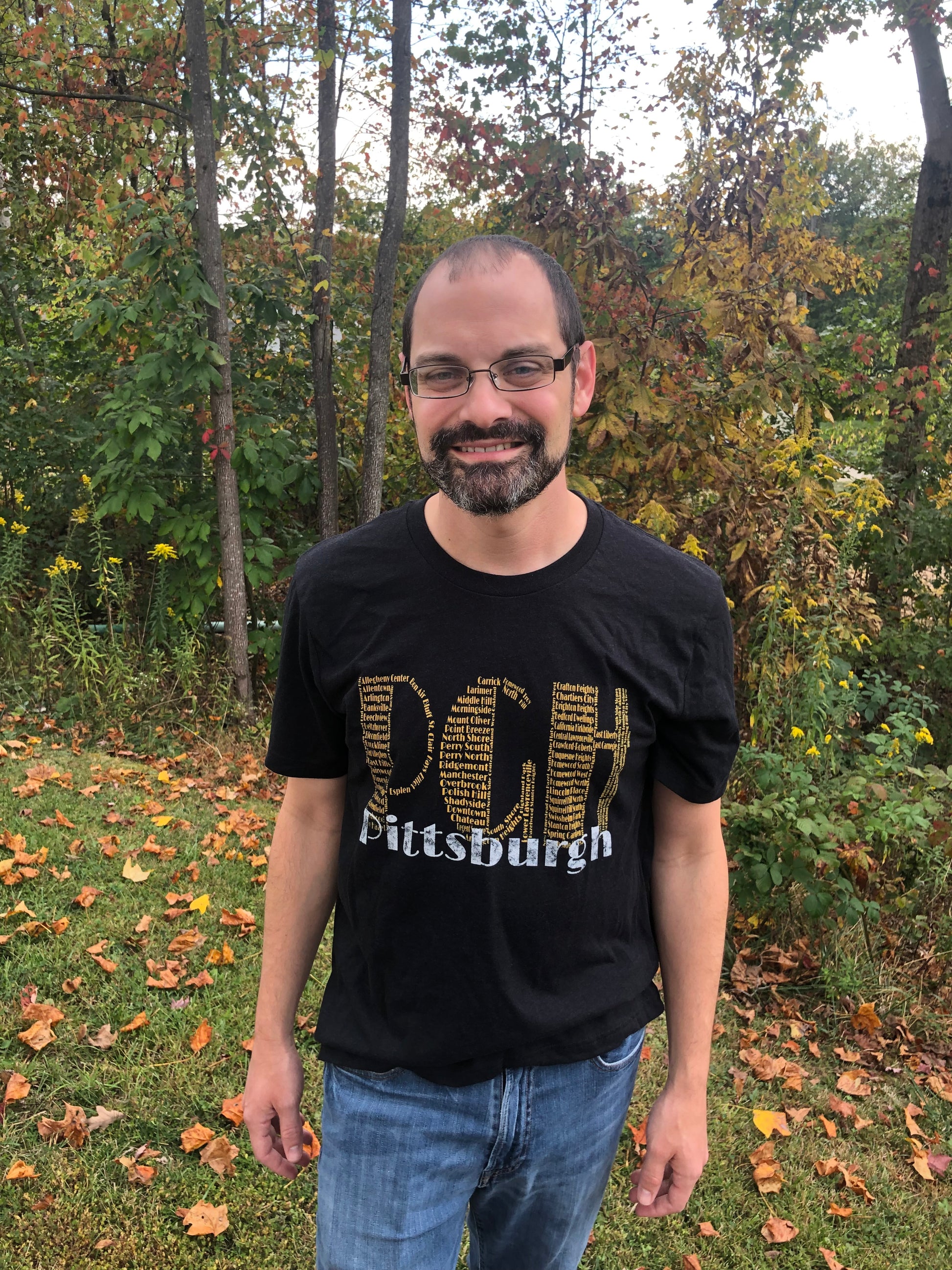 Man wearing a black t-shirt with 'Pittsburgh' text standing in a forested area with autumn leaves.