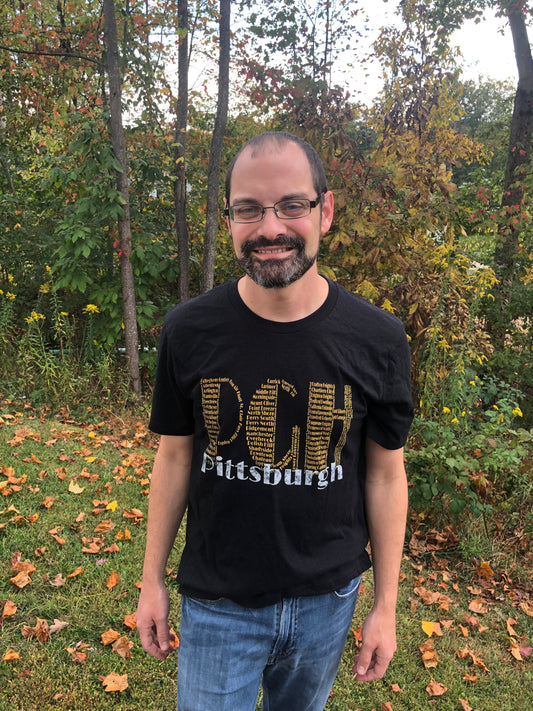 Man wearing a black t-shirt with 'Pittsburgh' text standing in a forested area with autumn leaves.
