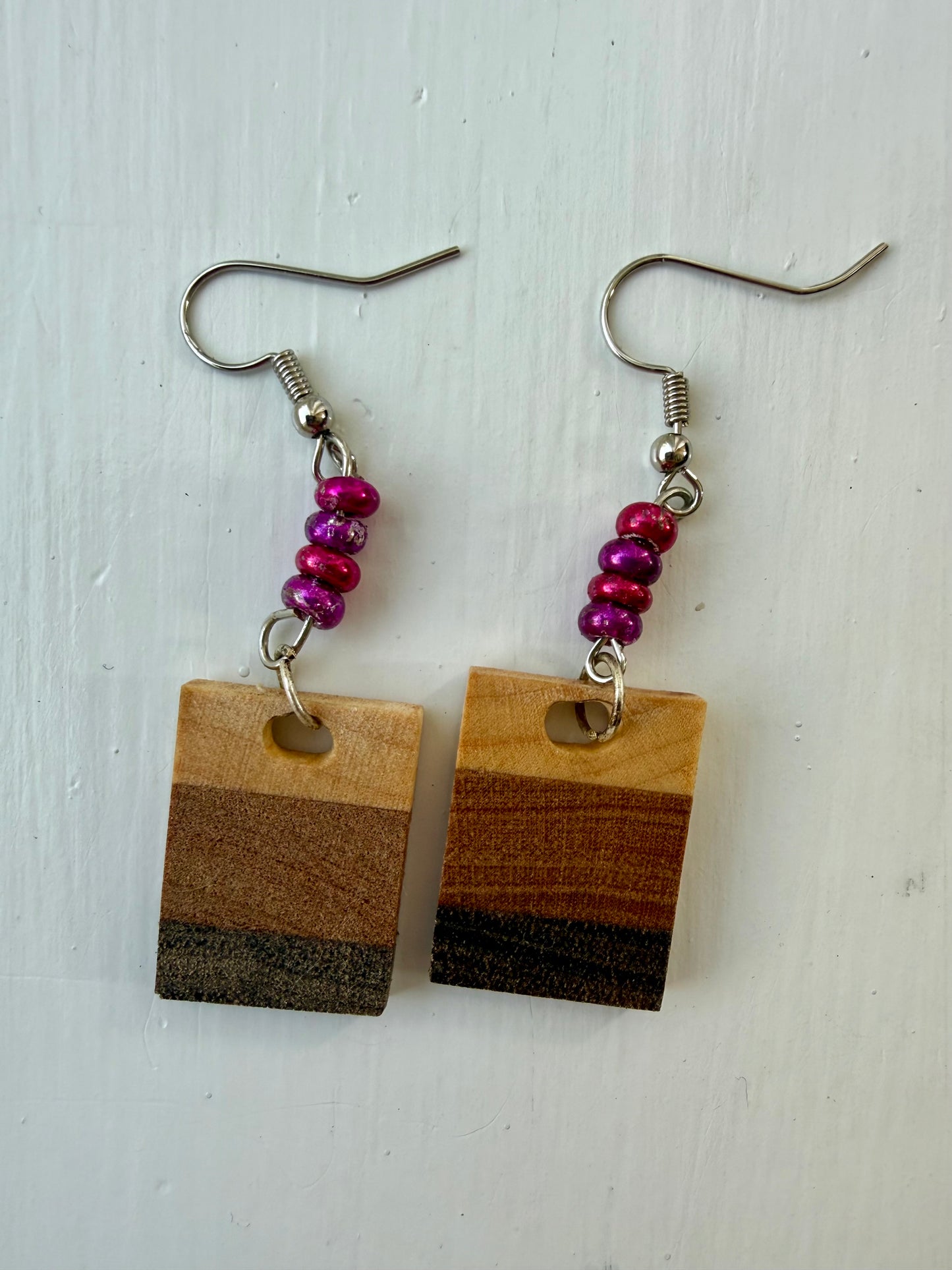 Two rectangular drop earrings featuring three horizontal wood-patterned stripes in light tan, medium brown, and dark charcoal. Each earring hangs from a silver-coated metal ear hook with small pink and purple glass bead accents.