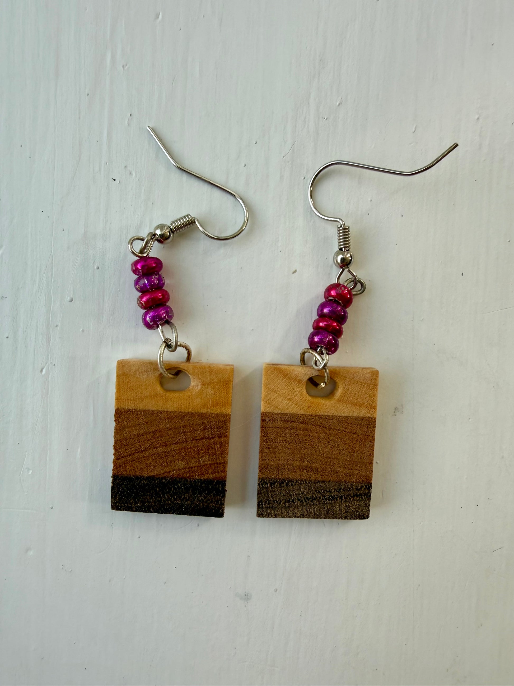 Two rectangular drop earrings featuring three horizontal wood-patterned stripes in light tan, medium brown, and dark charcoal. Each earring hangs from a silver-coated metal ear hook with small pink and purple glass bead accents.