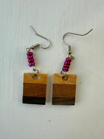 Two rectangular drop earrings featuring three horizontal wood-patterned stripes in light tan, medium brown, and dark charcoal. Each earring hangs from a silver-coated metal ear hook with small pink and purple glass bead accents.