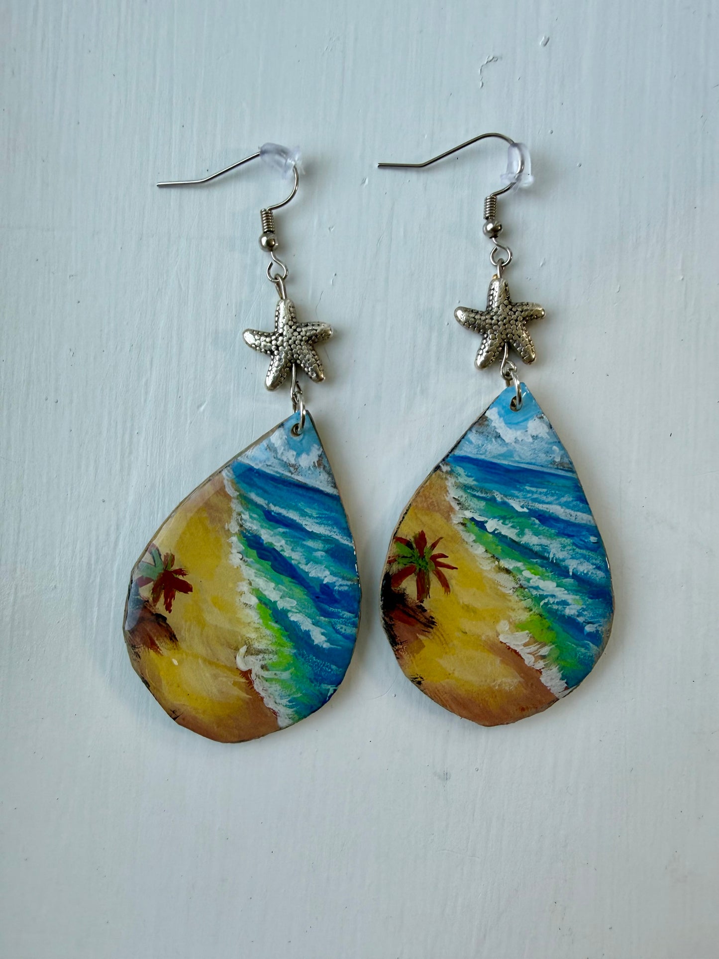 A pair of teardrop-shaped dangling earrings with painted beach scenes in blue, green, yellow, and brown, hanging from silver coated metal hooks with starfish charms.