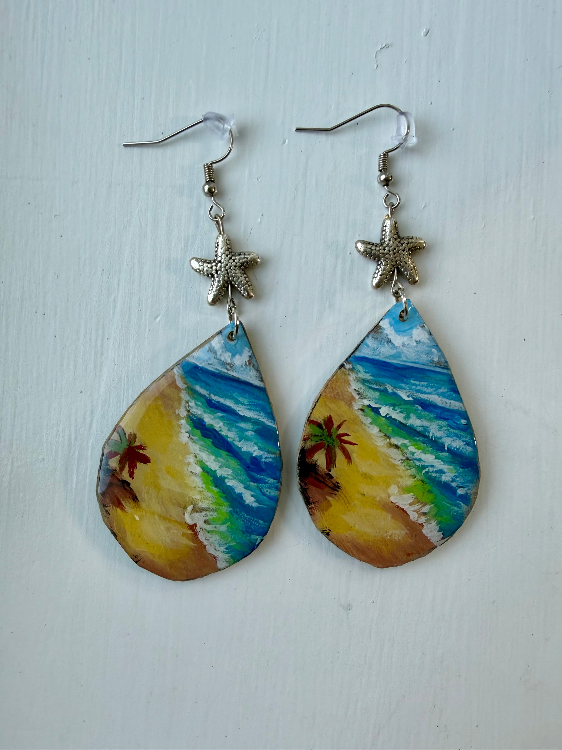 A pair of teardrop-shaped dangling earrings with painted beach scenes in blue, green, yellow, and brown, hanging from silver coated metal hooks with starfish charms.