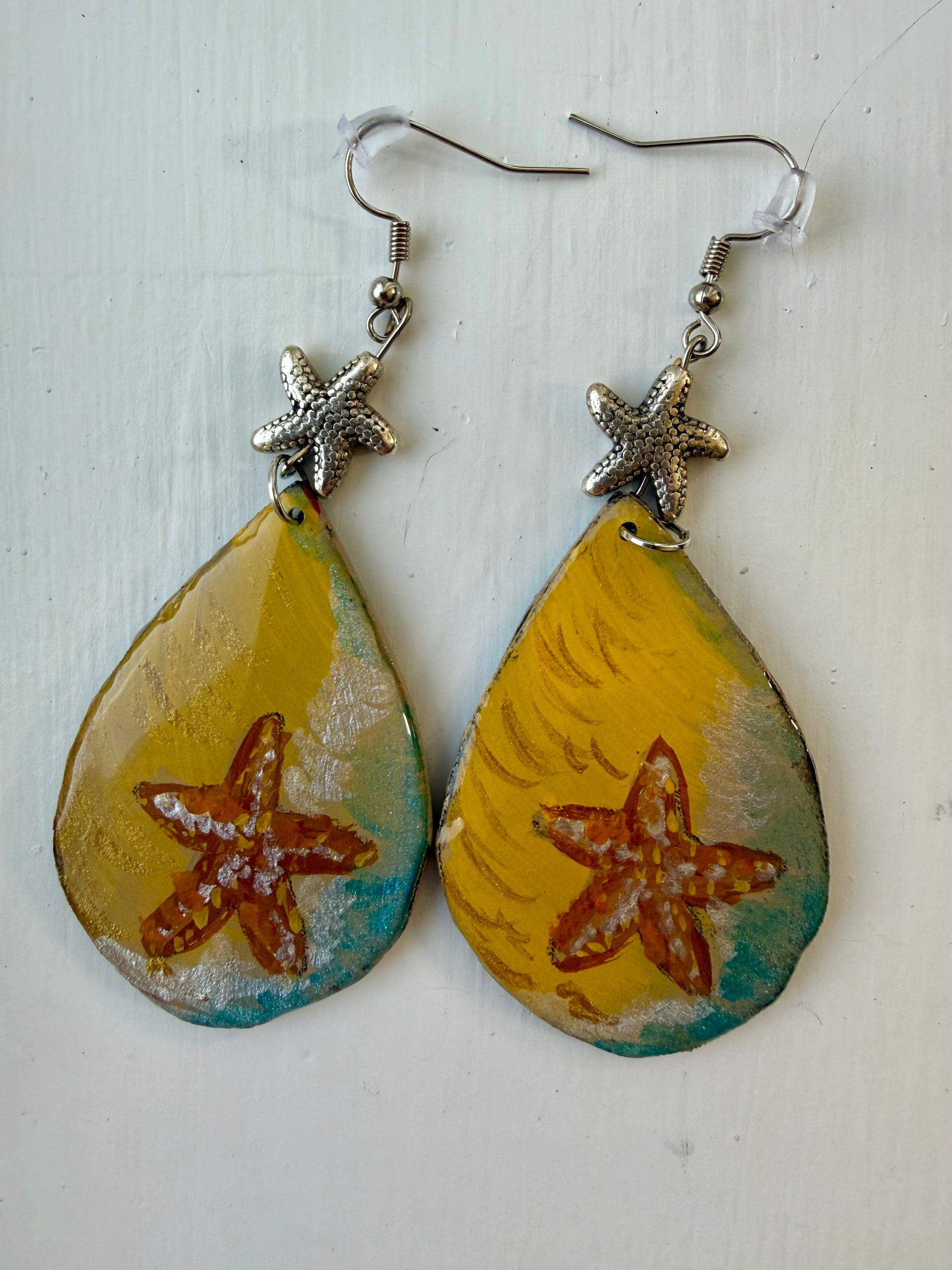 A pair of teardrop-shaped dangling earrings featuring a yellow and teal marbled design with a painted orange-brown starfish in the center, hanging from silver coated metal hooks with starfish charms.