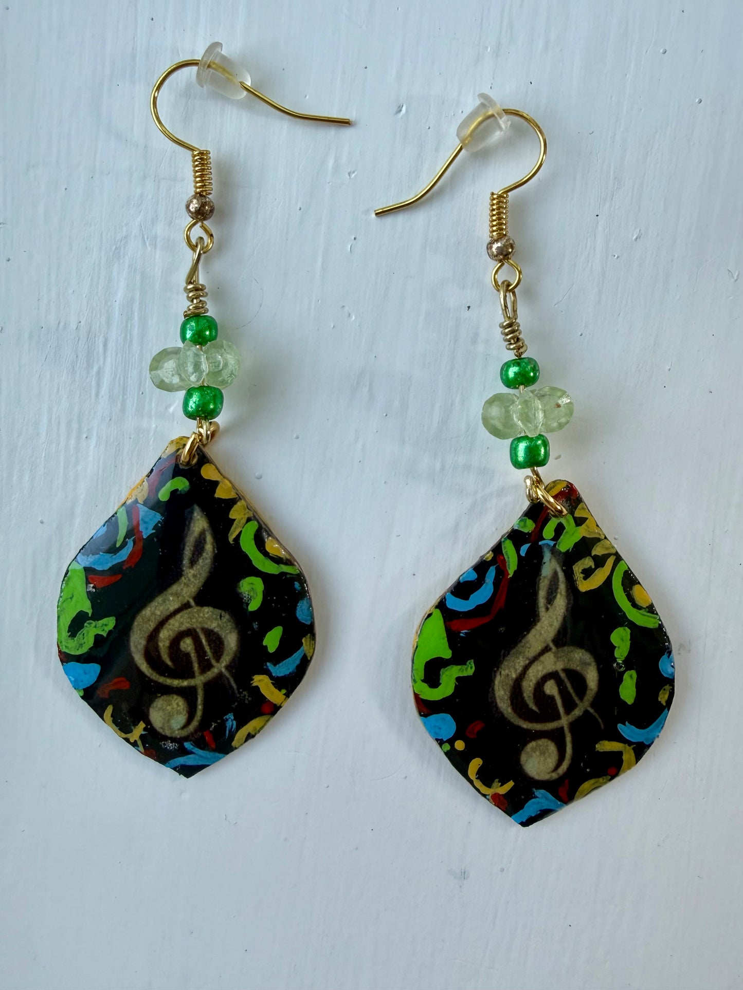 A pair of teardrop-shaped dangle earrings with gold-coated metal hooks and green beads, featuring a colorful abstract pattern with a gold treble clef on a black background, displayed on a white background.