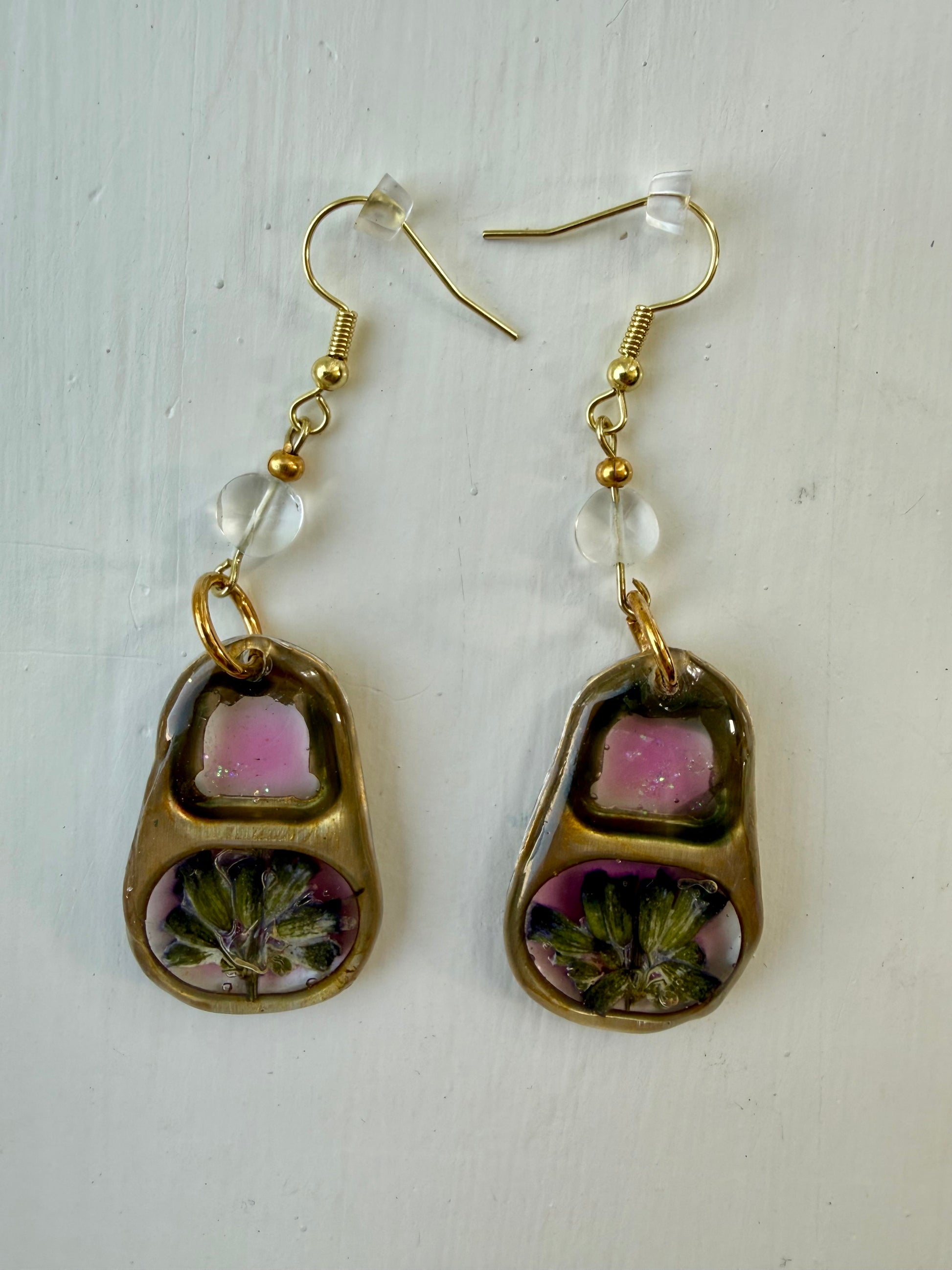 A pair of gold-tone dangle earrings with blue topaz beads and pink and floral inlay, displayed on a white textured background.