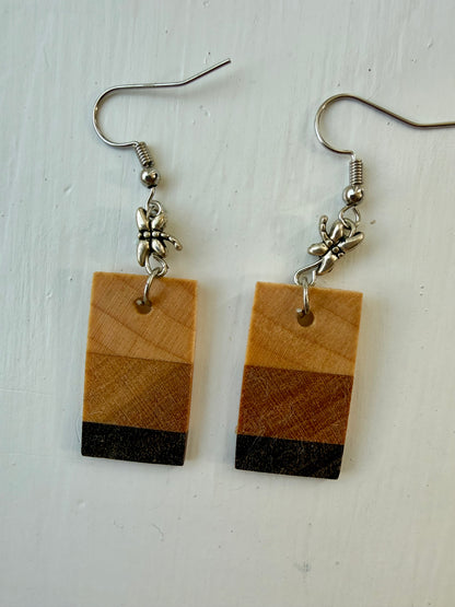 Two rectangular drop earrings featuring three horizontal wood-patterned stripes in light tan, medium brown, and dark charcoal. Each earring hangs from a silver-coated metal ear hook with a small silver-coated metal dragonfly bead.