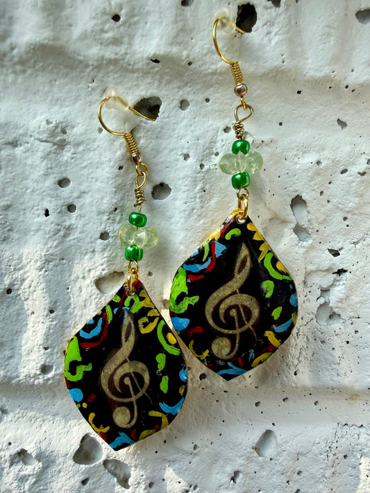 A pair of teardrop-shaped dangle earrings with gold-coated metal hooks and green beads, featuring a colorful abstract pattern with a gold treble clef on a black background, displayed against a textured white wall.