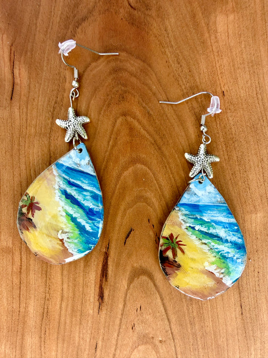 A pair of teardrop-shaped dangling earrings with painted beach scenes in blue, green, yellow, and brown, hanging from silver coated metal hooks with starfish charms on a wooden surface.