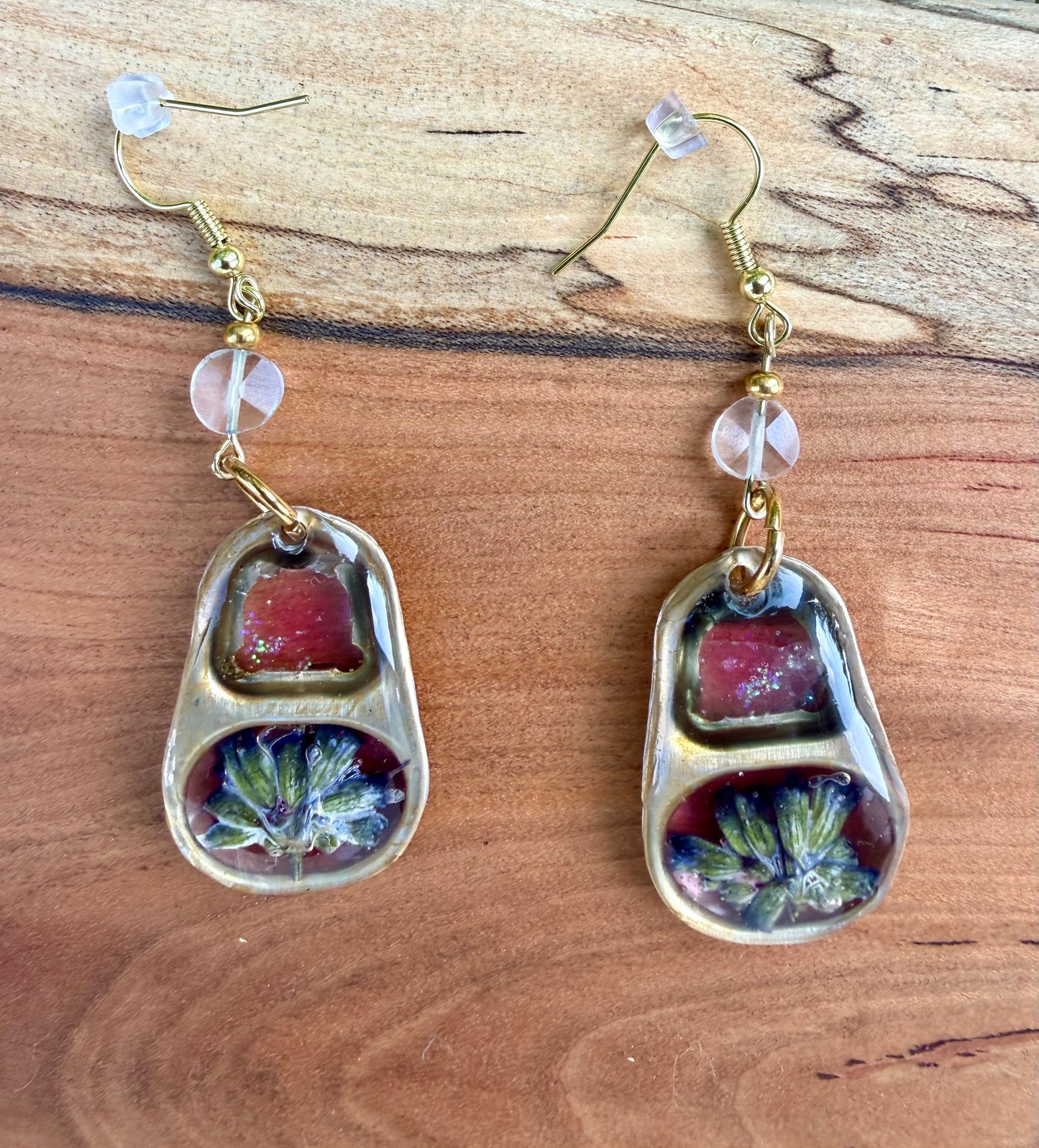 A pair of gold-tone dangle earrings with blue topaz beads and pink and floral inlay, displayed on a wooden background.