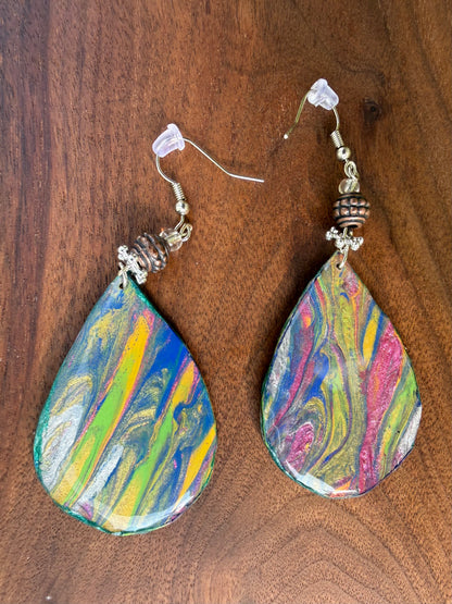 A pair of teardrop-shaped dangle earrings with colorful marbled paint designs in blue, green, yellow, pink, and white, hanging on silver coated metal hooks on a dark wood background.