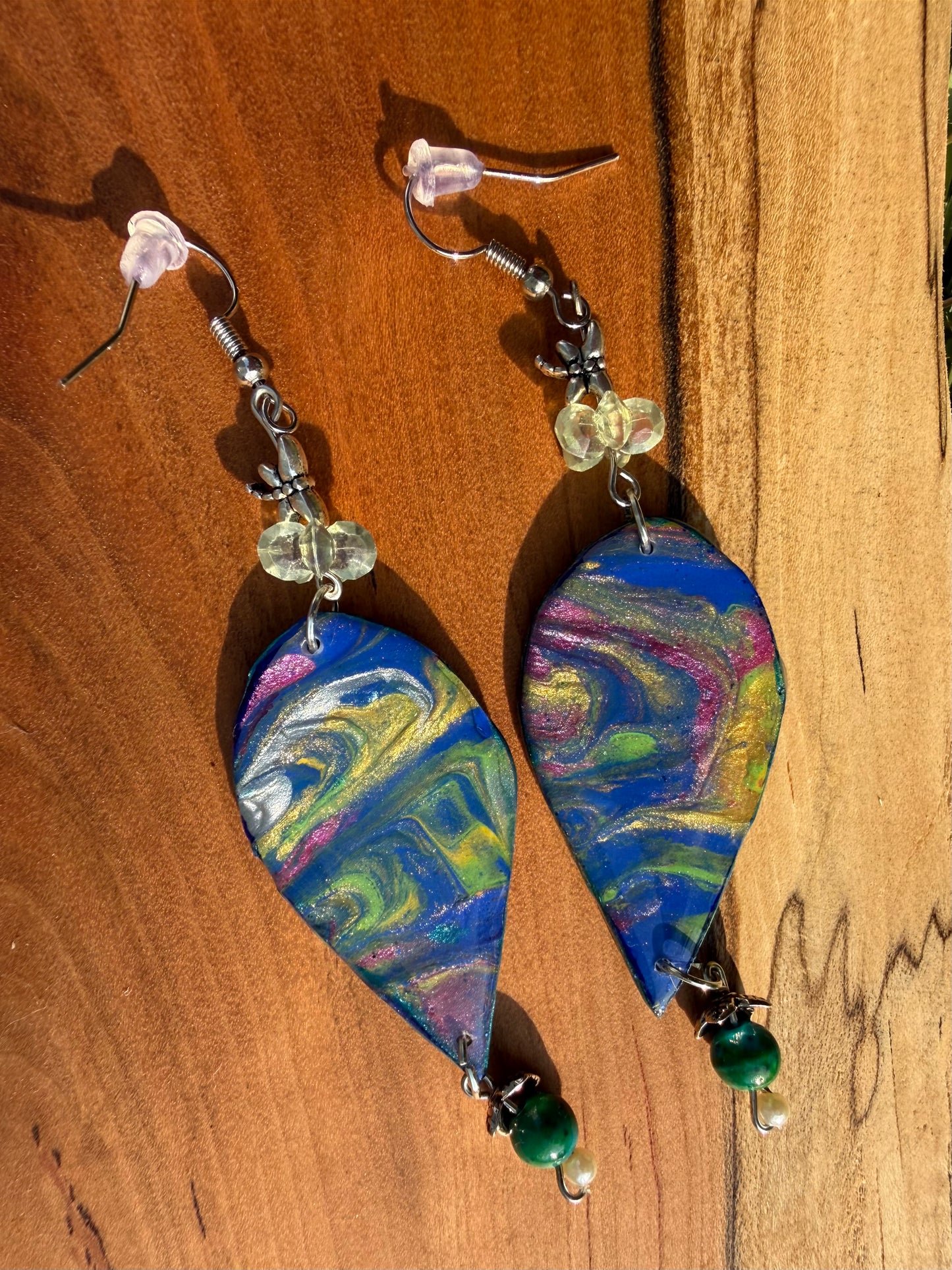 A pair of teardrop-shaped dangle earrings with colorful marbled paint designs in blue, green, yellow, pink, and white, hanging on silver coated metal hooks on a dark wood background.