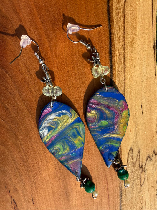 A pair of teardrop-shaped dangle earrings with colorful marbled paint designs in blue, green, yellow, pink, and white, hanging on silver coated metal hooks on a dark wood background.