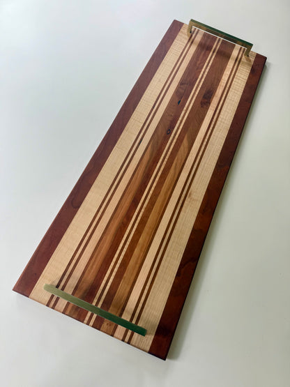 A rectangular wooden serving board with a striped pattern of light and dark wood tones, featuring brass colored handles