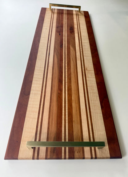 A rectangular wooden serving board with a striped pattern of light and dark wood tones, featuring brass colored handles